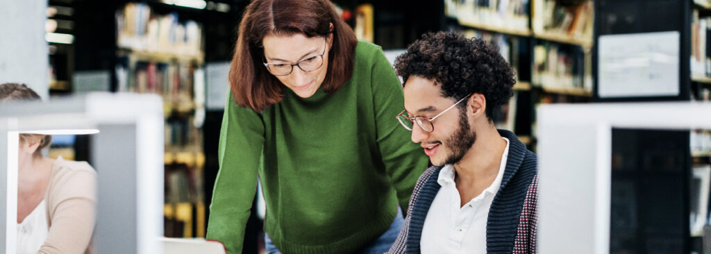 A woman and a man are working at a desk. The woman is standing behind the man who is sitting. They're both looking at a laptop.