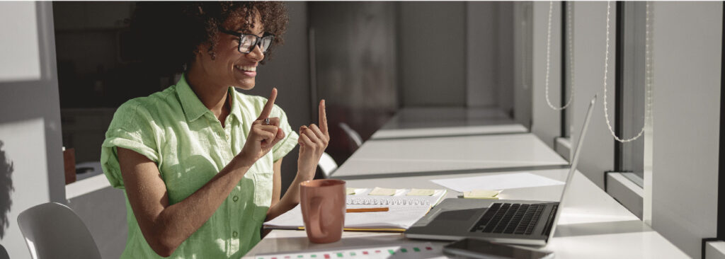 Woman sitting at her desk, working at her laptop, and gesturing toward the screen with her hands, one held in front of the other with pointer fingers raised