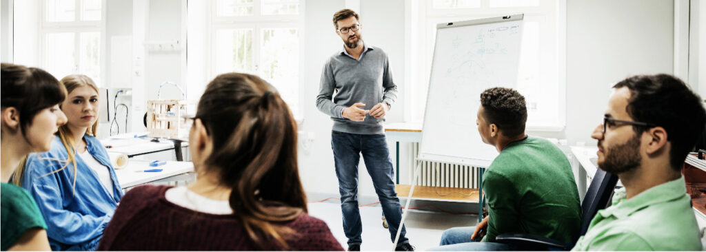 Man standing at the front of a classroom talking to a small group of students with a whiteboard beside him
