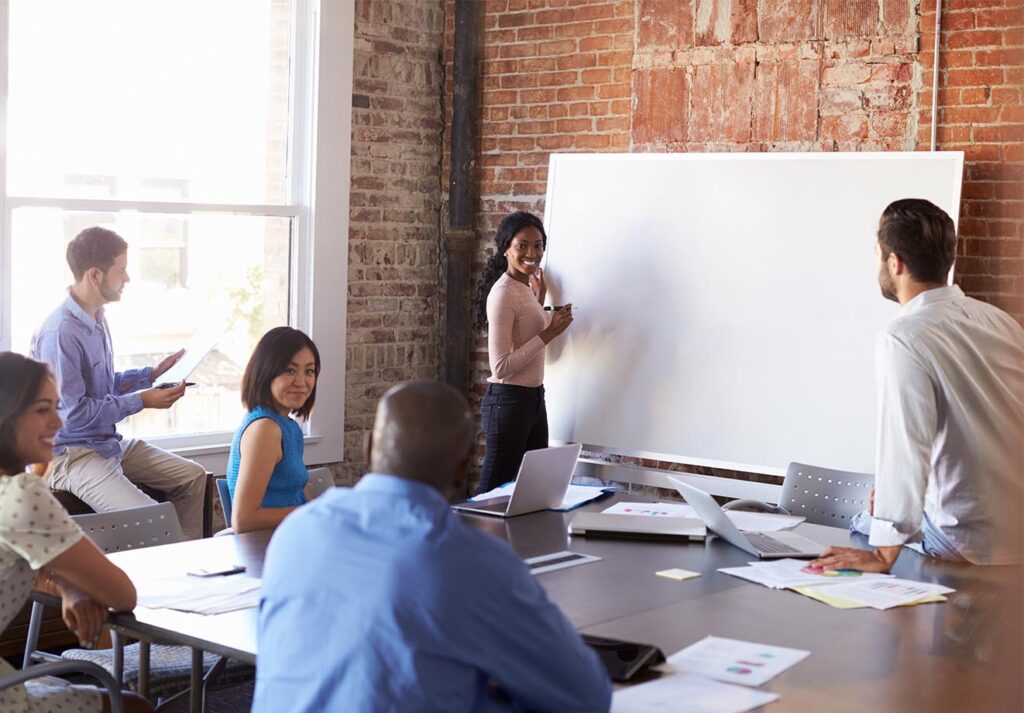 Six people sit in a room with a big window and a brick wall, taking ideas and writing them on a whiteboard